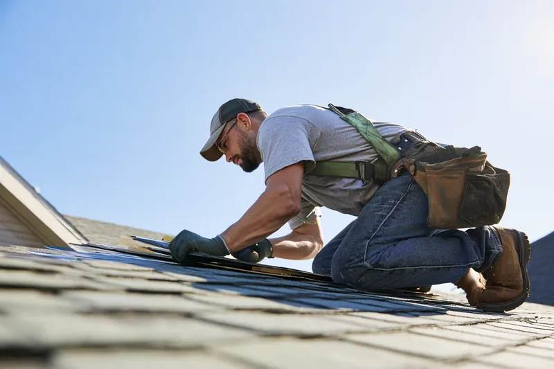 Roofer in his 30s replacing asphalt shingles on a suburban house roof under a clear blue sky