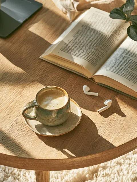 Top-down flat lay of a ceramic mug, an open paperback, wireless earbuds and a small houseplant on a light oak coffee table