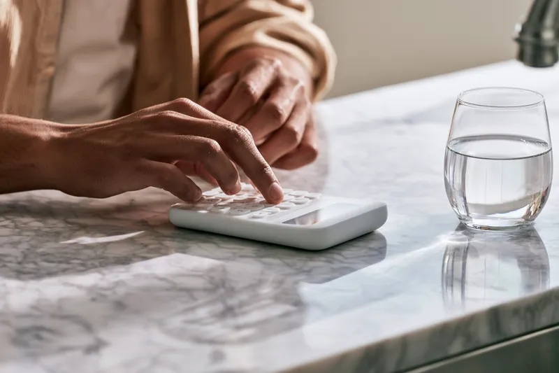 Hands of a man in his thirties tapping numbers into a sleek white calculator on a marble kitchen counter while estimating a Hidden Meadow Lending loan payment