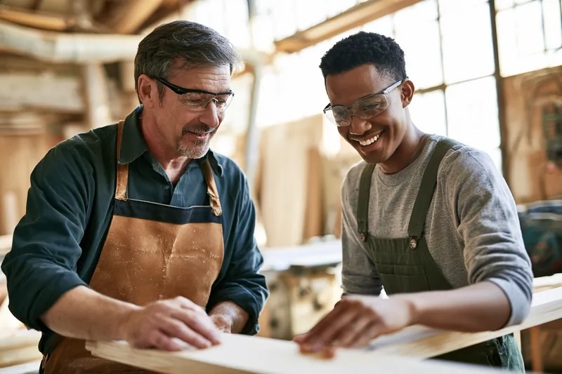 Two carpenters, one in his forties and one in his twenties, checking a freshly cut board in a woodworking shop